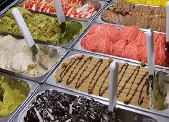 A display case filled with various flavors of gelato, including green, brown, pink, white, and yellow, some garnished with nuts, chocolate drizzle, or fruit, with white plastic serving scoops in several trays.