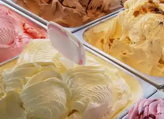 Several trays of colorful ice cream in a display case, including flavors like vanilla, strawberry, and chocolate. A plastic spatula rests in the creamy, swirled surface of the vanilla ice cream.