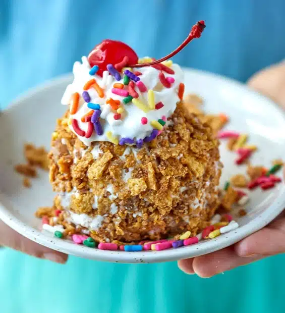 A person holds a plate with a scoop of fried ice cream topped with whipped cream, colorful sprinkles, and a cherry. Some sprinkles are scattered on the plate.