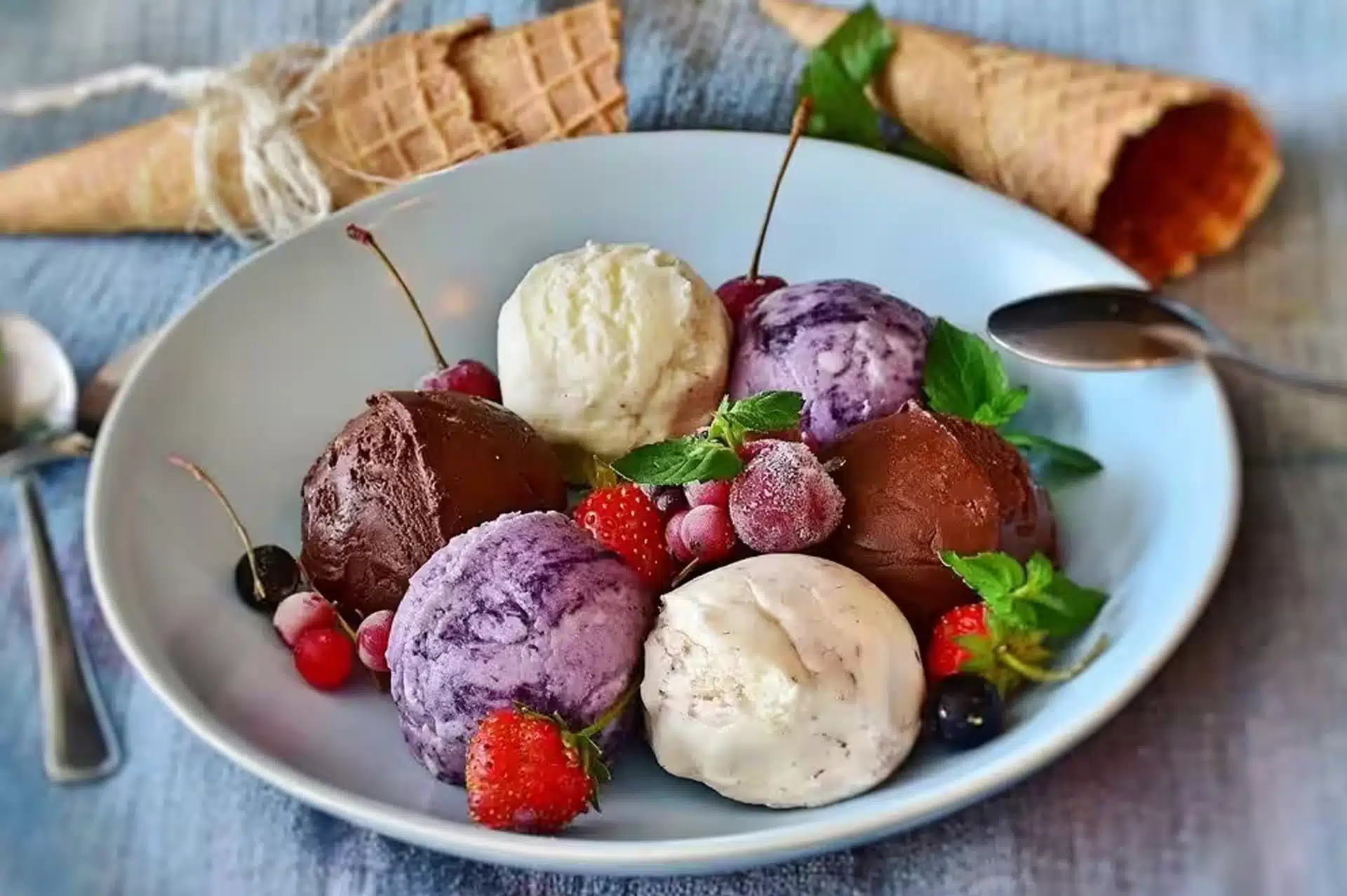 A white plate holds assorted ice cream scoops in chocolate, vanilla, and berry flavors, garnished with fresh berries and mint. Two empty waffle cones and spoons sit on a blue cloth background.