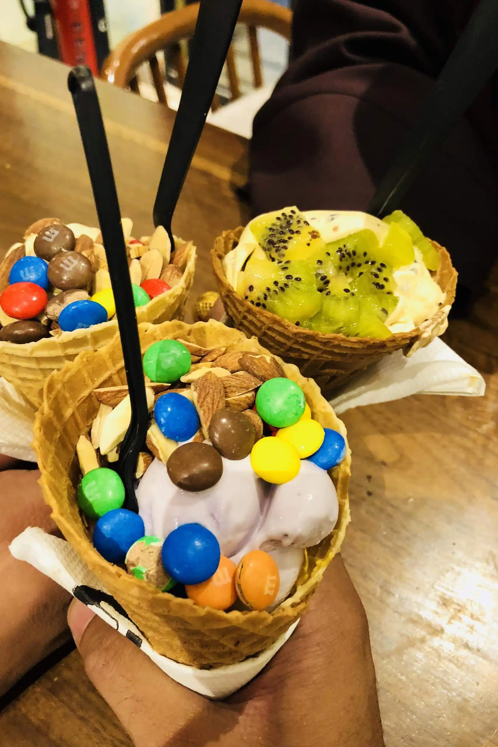 Three hands hold waffle cones filled with colorful ice cream, topped with M&Ms, chopped almonds, and sliced kiwi. Each cone has a black spoon and is wrapped in a napkin, set against a wooden table background.