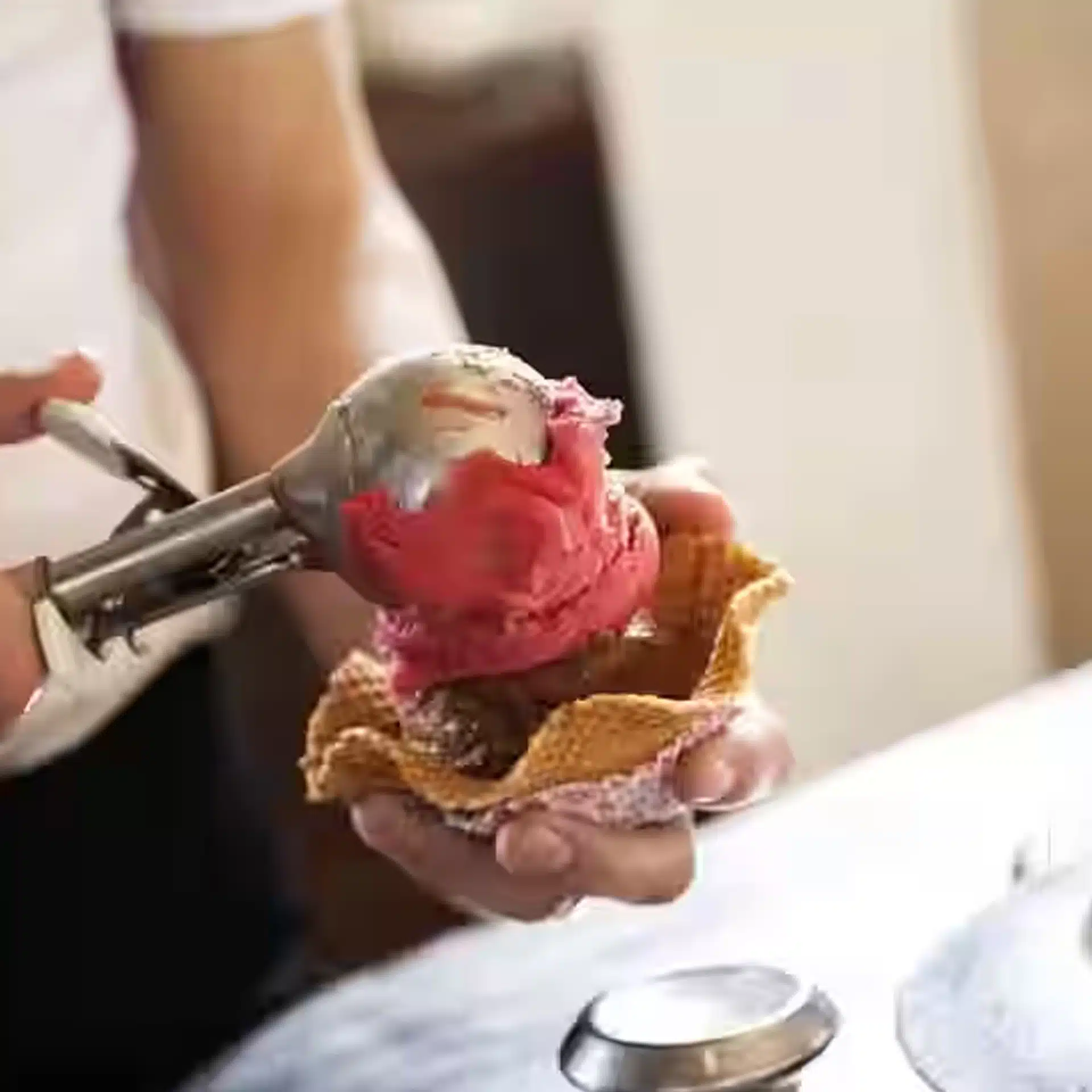 A person uses an ice cream scoop to serve a scoop of pink ice cream onto a waffle bowl already holding a scoop of chocolate ice cream.