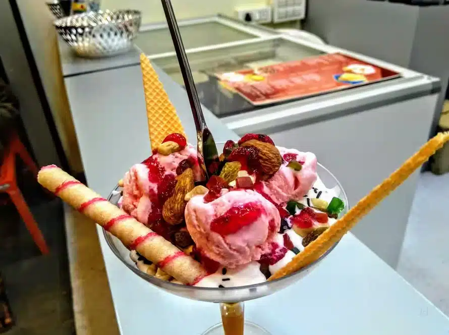 A glass dessert cup filled with scoops of pink ice cream, topped with nuts, berries, and syrup, garnished with wafer sticks and a triangular wafer, with a spoon placed upright in the center.