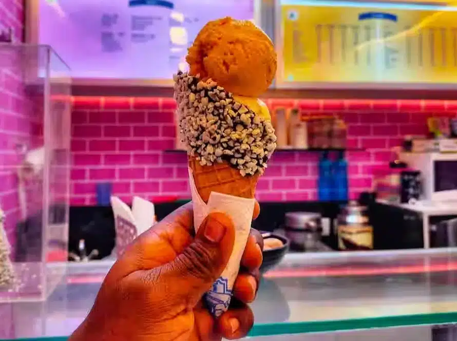 A hand holds an ice cream cone with two scoops and chocolate sprinkles, against a backdrop of a pink tiled wall and a brightly lit ice cream shop counter.